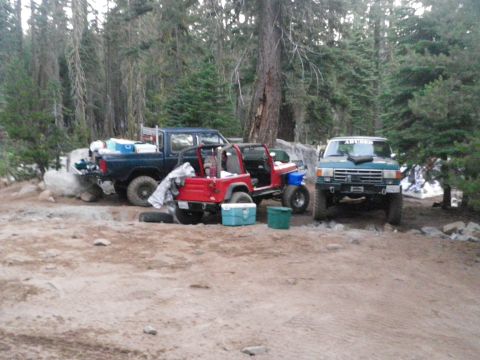 1991 Ford Bronco - Multiple off-road vehicles are parked in a forest clearing with camping gear.