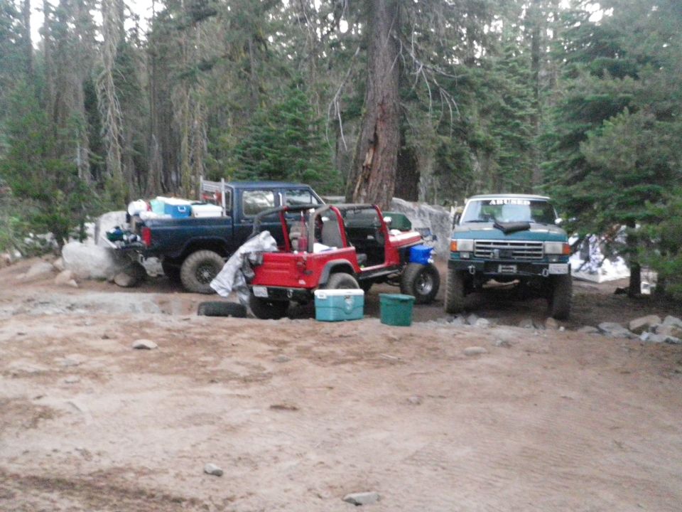 Blue pickup truck, red Jeep, and green SUV parked in a wooded area. Coolers and camping gear are visible
