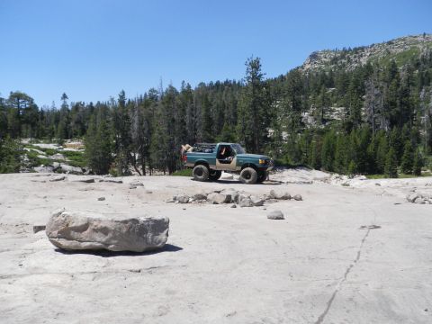1991 Ford Bronco - A modified Ford Bronco sits on a rocky terrain with a forest and mountain backdrop under a clear blue sky.