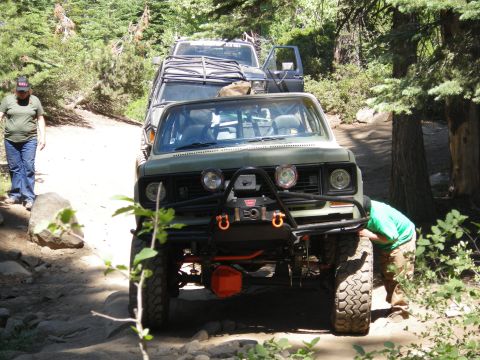 1991 Ford Bronco - Two off-road vehicles are stopped on a dirt trail, with one person working on the front of the lead vehicle.