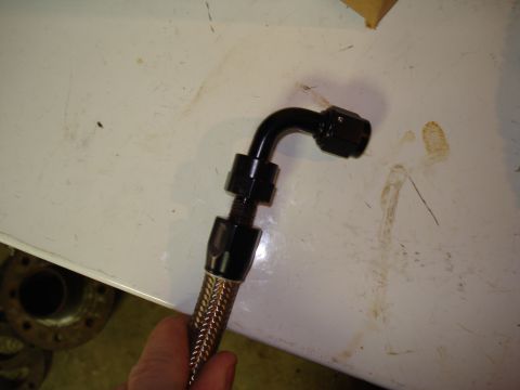 1984 Ford Bronco - A hand holds a braided stainless steel line assembled with a black anodized straight AN hose end and a 90-degree elbow fitting, held agains…