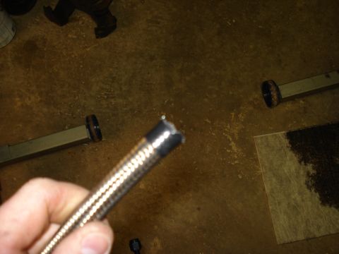 1984 Ford Bronco - A hand holds a freshly cut length of braided stainless steel hose, showing the taped cut end with a few frayed strands, during AN fuel line…