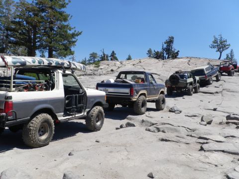 1991 Ford Bronco - A convoy of modified off-road vehicles, including a 1991 Ford Bronco, navigates a challenging rocky path under a clear blue sky.