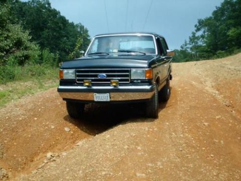 1989 Ford Bronco - This 1989 Ford Bronco is captured from the front on a dusty, unpaved road surrounded by trees.