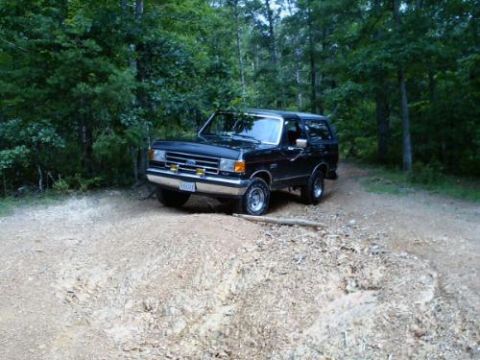 1989 Ford Bronco - Black Ford Bronco on a dirt road surrounded by trees.