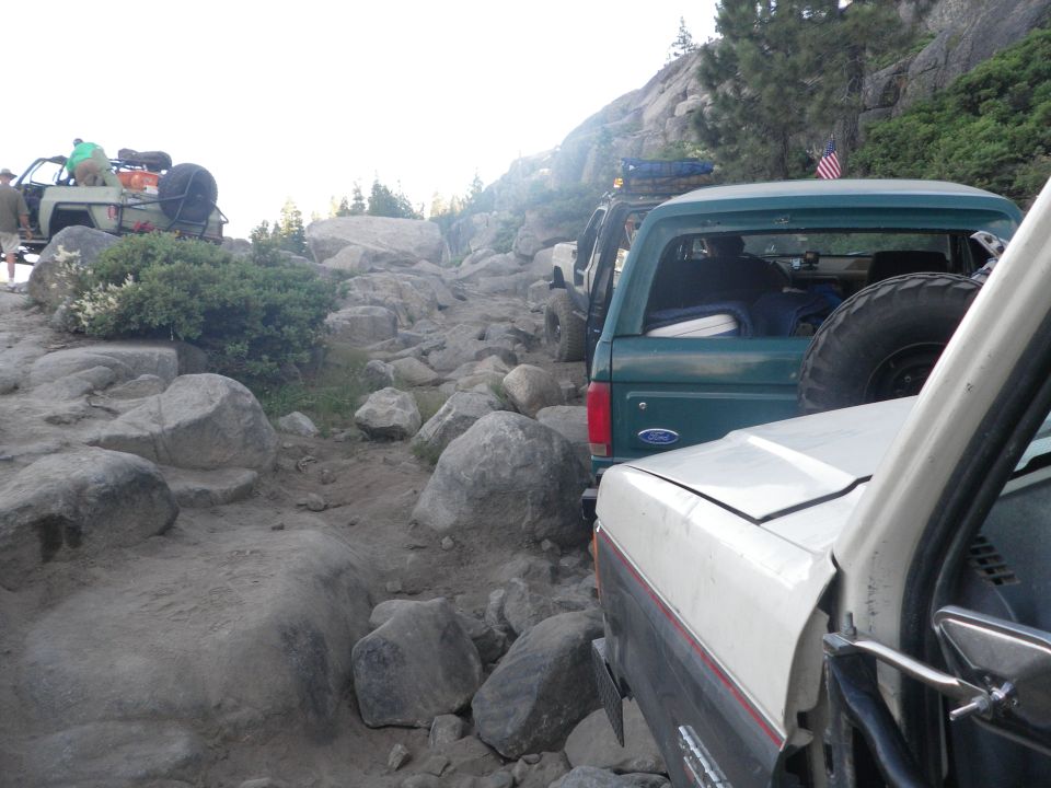 Line of off-road vehicles, including a 1991 Ford Bronco, navigating a rocky mountain trail