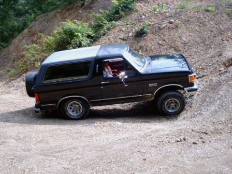 1989 Ford Bronco - A black Ford Bronco navigates a dirt road with a wooded hillside in the background.