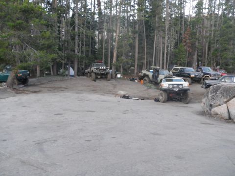 1991 Ford Bronco - Several off-road vehicles, including a white 1991 Ford Bronco, are parked in a campsite surrounded by trees.