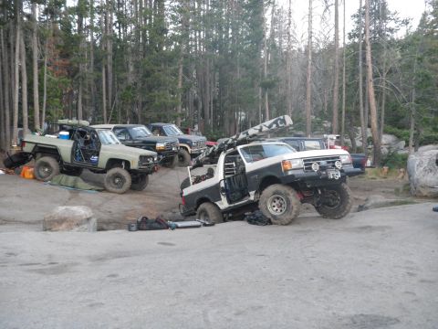 1991 Ford Bronco - Several modified off-road vehicles, including a 1991 Ford Bronco, are parked in a forest clearing.