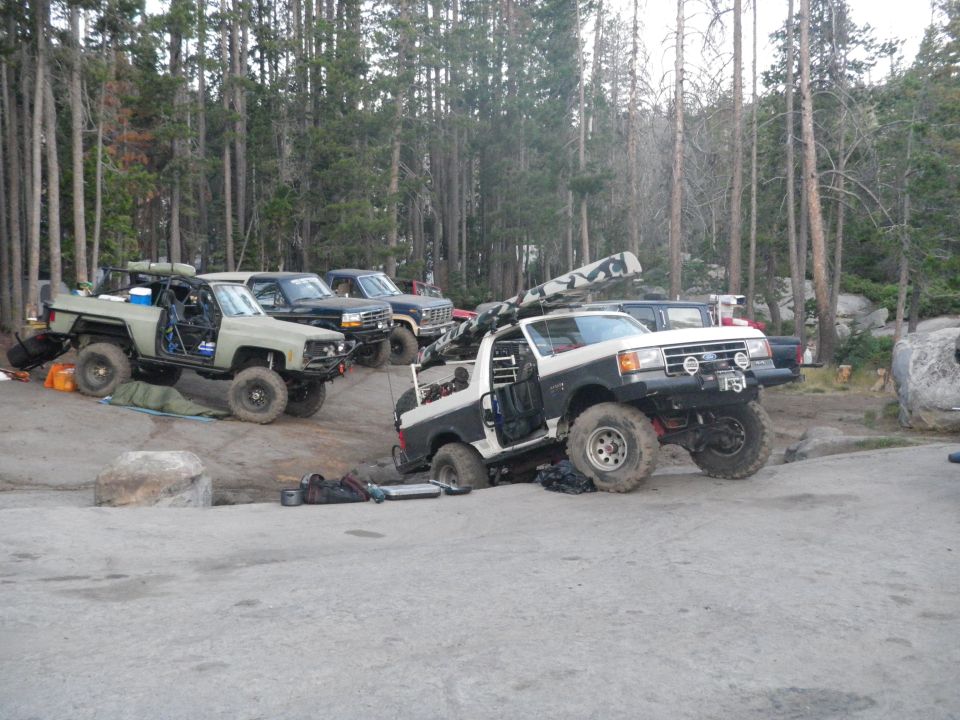 Modified 1991 Ford Bronco with off-road tires and roll cage parked with other trucks in a wooded area
