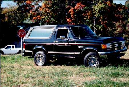 1989 Ford Bronco - A black 1989 Ford Bronco is seen parked on a grassy area with a stop sign and a white pickup truck in the background.