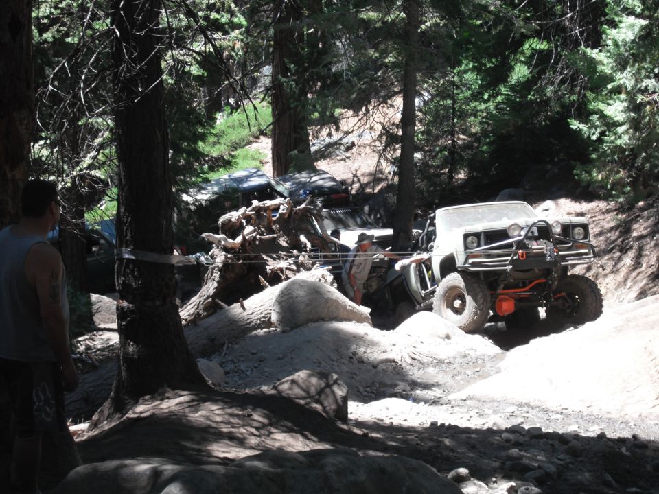 Off-road vehicle with winch and roll cage navigating a rocky, wooded trail. Another vehicle is visible behind it