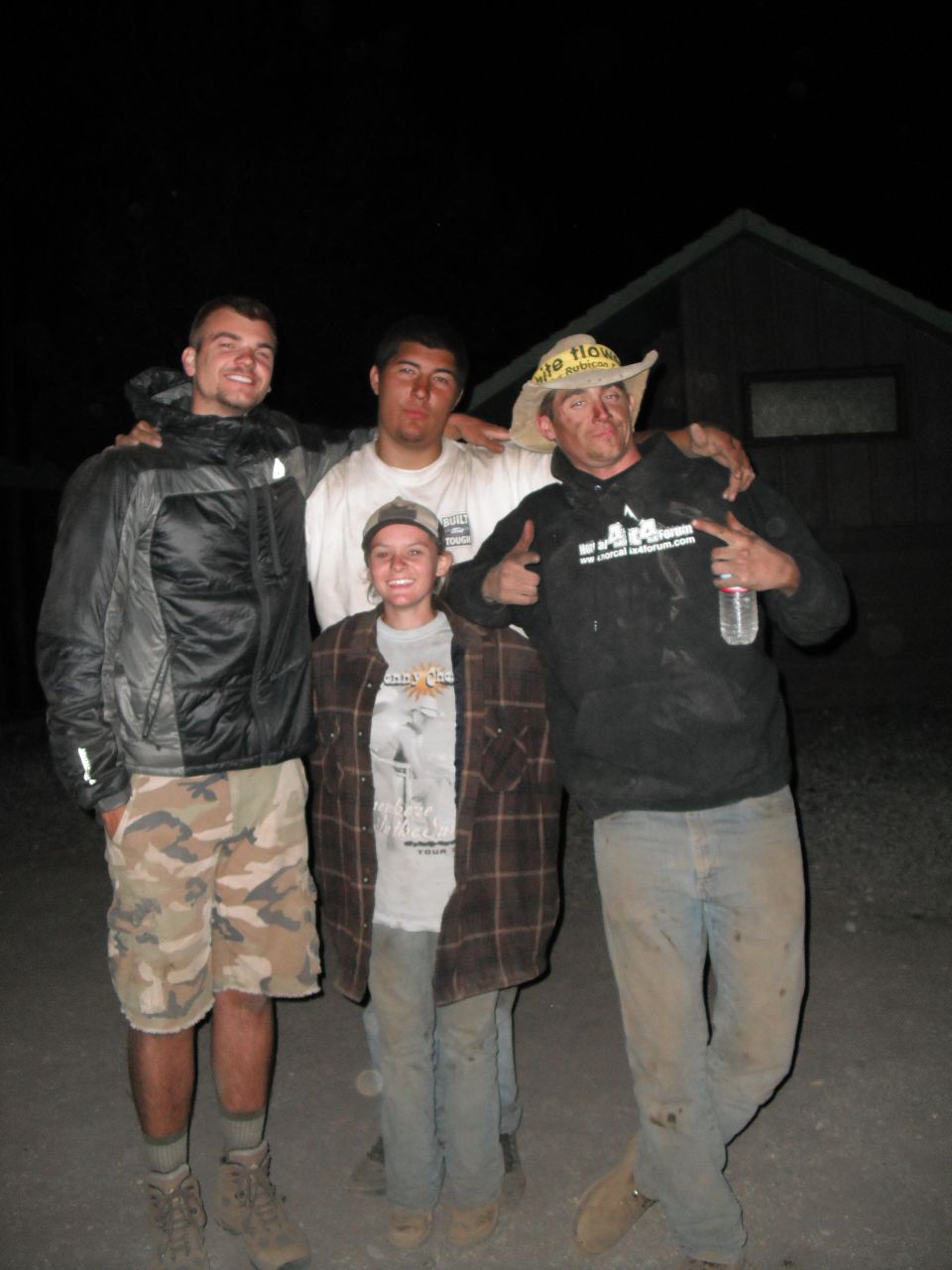 Four people stand in front of a dark building at night. The person on the far right wears a cowboy hat and a black hoodie