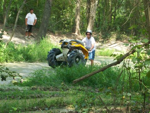2008 Bombardier Outlander - An ATV navigates a muddy trail in a forest with a person riding it and another standing nearby.