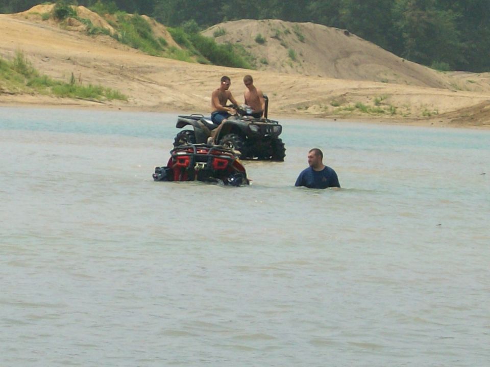 Two ATVs submerged in a sand pit pond with riders and a man standing in water