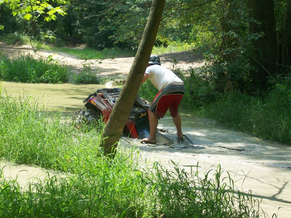 Red Can-Am ATV stuck in a muddy creek as a man works to free it on a wooded trail