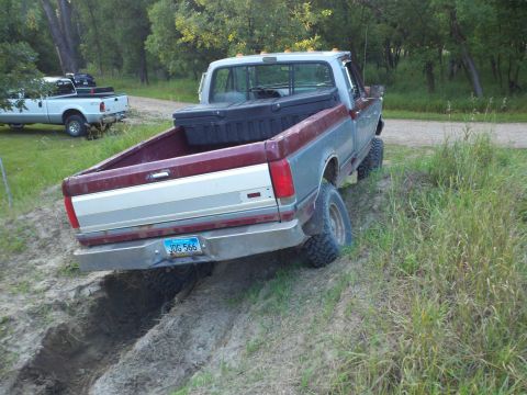 1988 Ford F-150 - A red and gray Ford pickup truck is stuck in a ditch with another white pickup truck in the background.