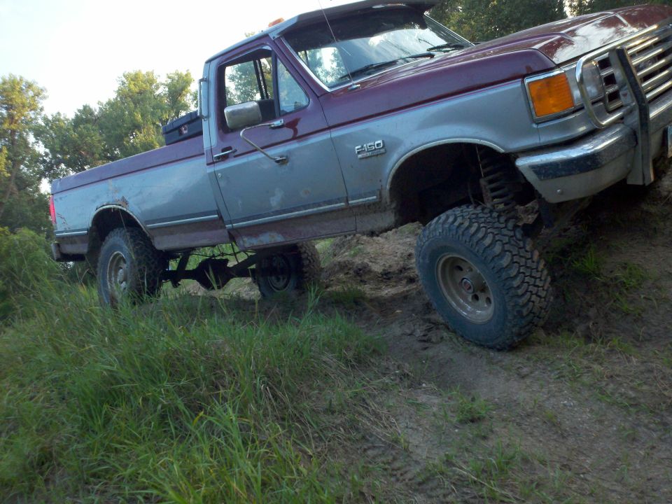 1988 Ford F-150 pickup truck with two-tone paint, lifted suspension, and off-road tires driving on a dirt path with grass