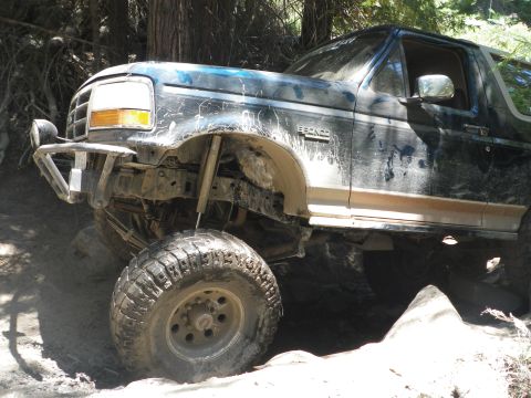 1991 Ford Bronco - This lifted SUV, likely a 1992 model, is shown tackling a challenging off-road trail with significant mud and rock obstacles.