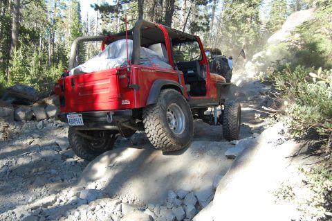1993 Jeep Wrangler - A red 1993 Jeep Wrangler tackles a challenging rocky trail, with another vehicle visible in the background kicking up dust.