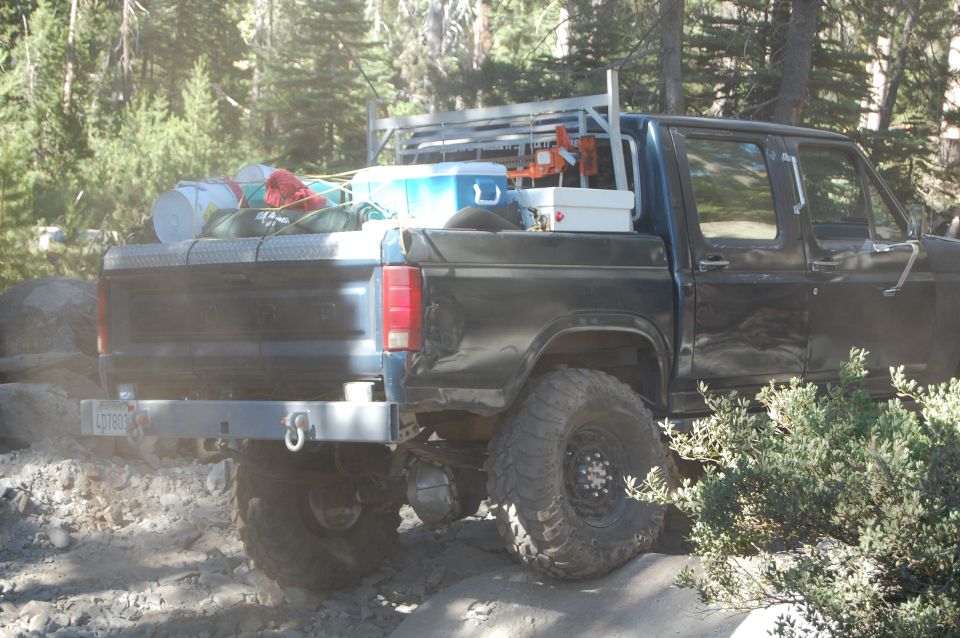 Rear view of a black pickup truck with a rack in the bed, loaded with camping gear and a cooler. License plate reads LGT803