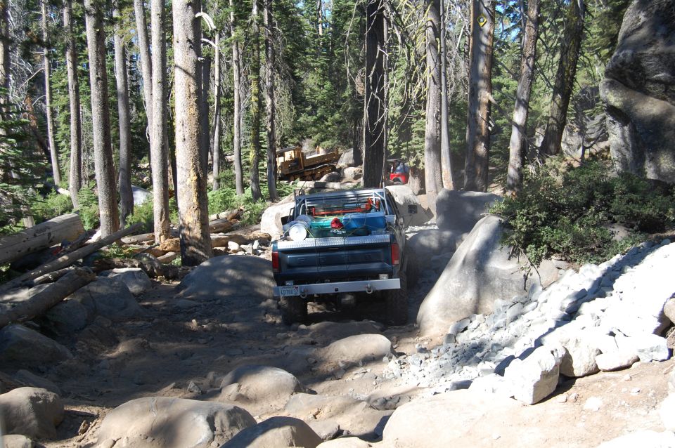 Pickup truck navigating a boulder-strewn forest trail. Another vehicle is visible further up the path
