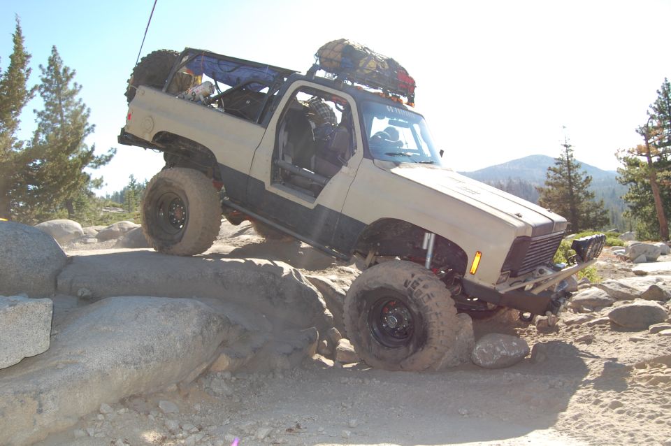 Tan off-road SUV with large tires navigating rocks, doors removed, spare tire on back, roof rack with gear