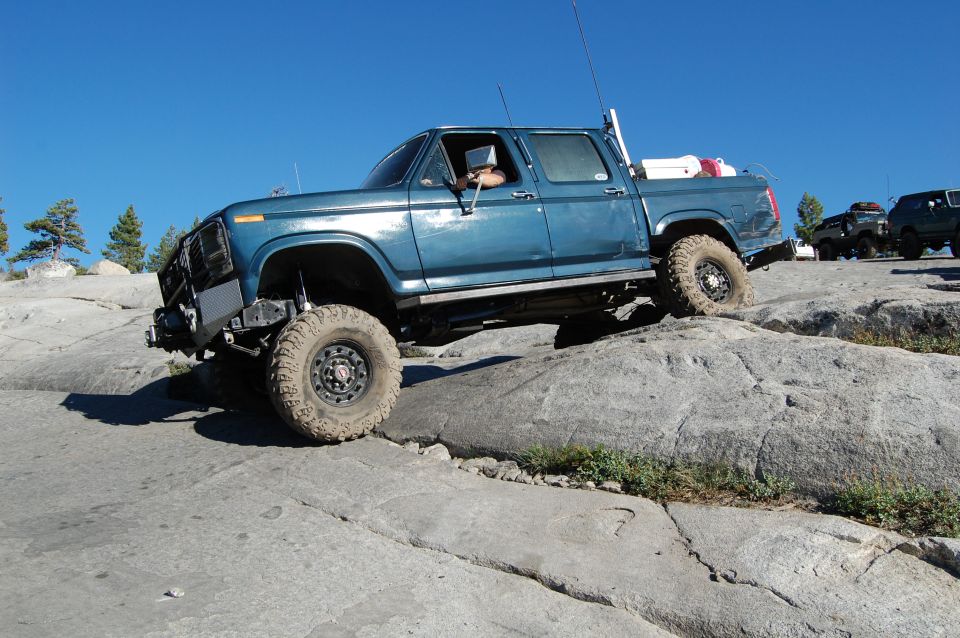Blue pickup truck with off-road tires climbing rocks, aftermarket bumper, driver visible