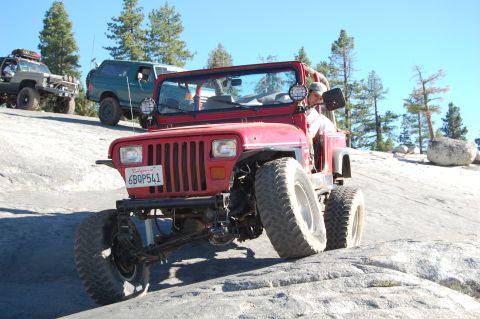 1993 Jeep Wrangler - A red Jeep Wrangler navigates a rocky terrain with other off-road vehicles in the background under a clear blue sky.