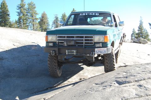 1993 Jeep Wrangler - This green truck, modified for off-road use, navigates a rocky terrain under a clear blue sky.