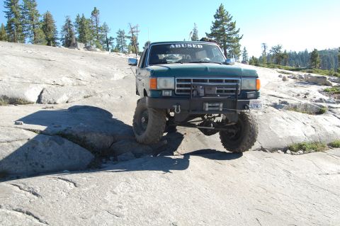 1993 Jeep Wrangler - A modified green truck navigates a rocky, off-road terrain under a clear blue sky.