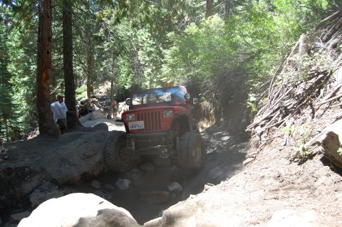 1993 Jeep Wrangler - A red Jeep Wrangler is shown tackling a challenging off-road trail with steep inclines and large rocks.