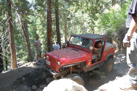 1993 Jeep Wrangler - A red 1993 Jeep Wrangler is driven on a challenging off-road path with trees and rocks surrounding it.