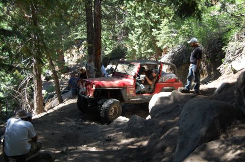 1993 Jeep Wrangler - A red Jeep Wrangler is shown off-roading on a steep, rocky path surrounded by trees.
