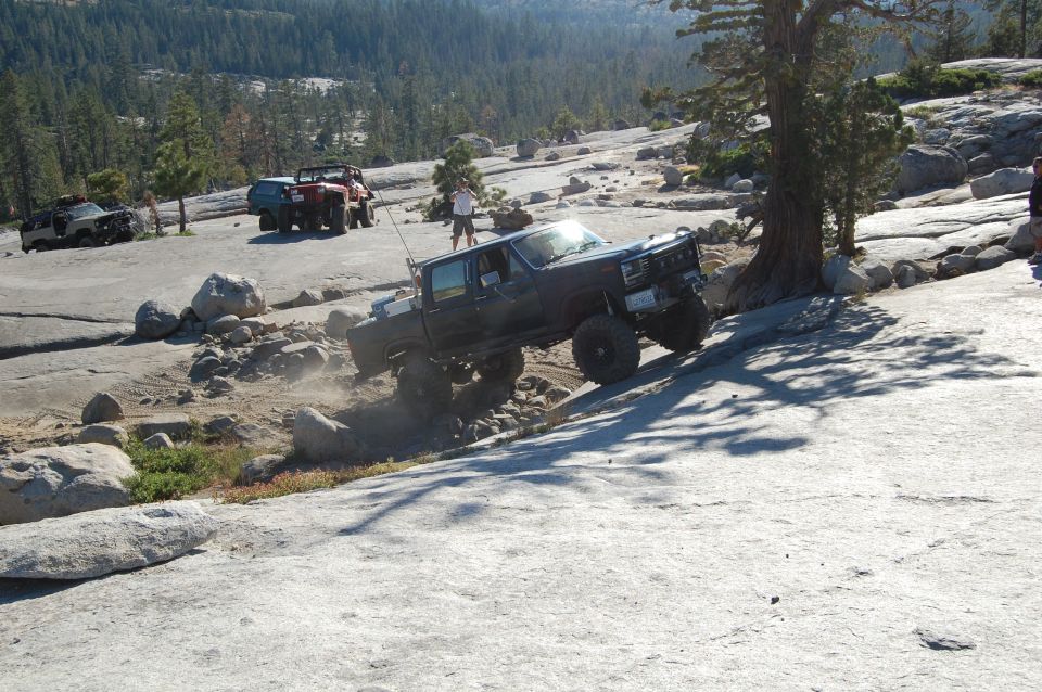 Dark pickup truck with large tires climbing a rocky incline, front-right wheel elevated
