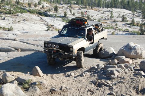 1993 Jeep Wrangler - A heavily modified pickup truck drives over large rocks on a sunny day.