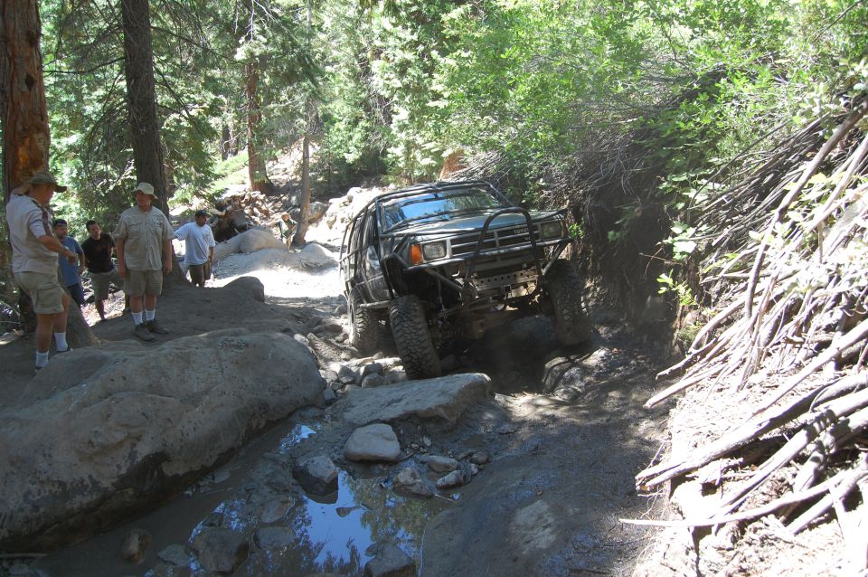 Off-road vehicle navigating a rocky, wooded trail with people watching