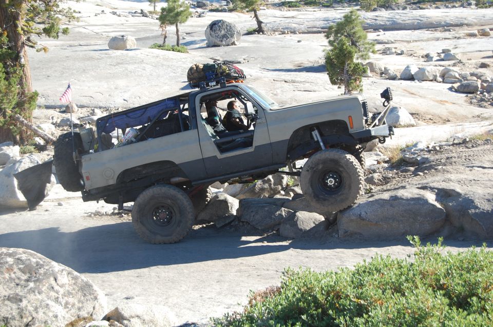 Tan and grey off-road SUV with large tires, driving over rocks. An American flag is mounted on the rear