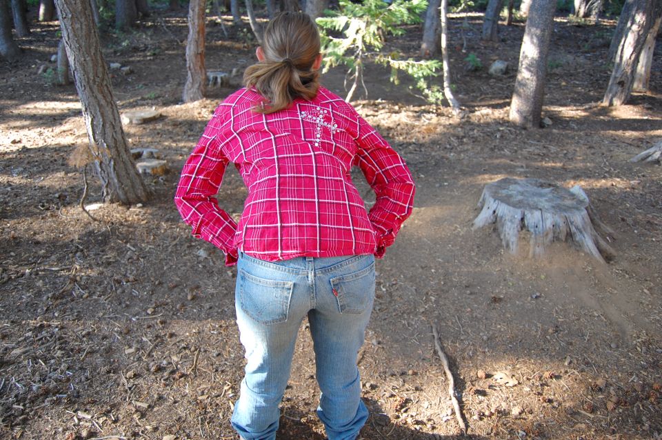 Person from behind wearing a plaid shirt and jeans, standing in a wooded area with trees and a tree stump