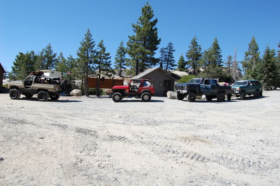 Four off-road vehicles parked in a dirt lot with trees and buildings in the background