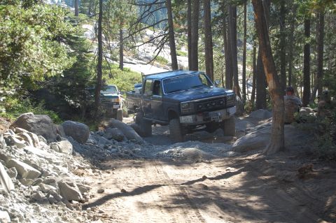 1993 Jeep Wrangler - Two trucks navigate a challenging, rocky path through a wooded area.
