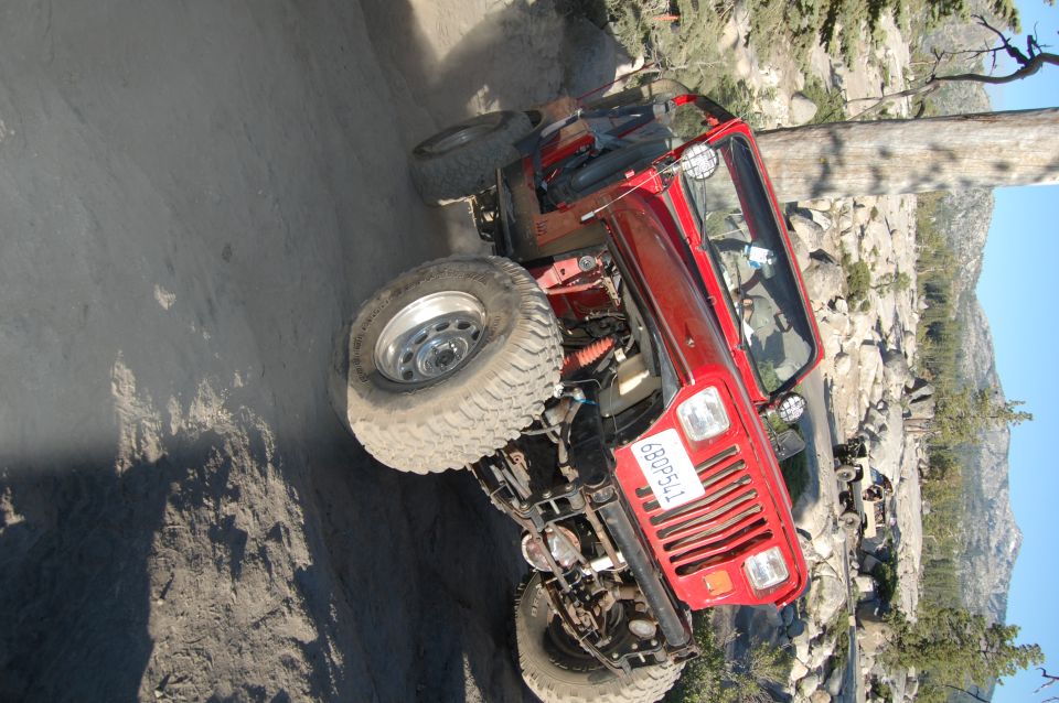 Red 1993 Jeep Wrangler on a rocky trail, front grille and tires visible. Another vehicle is parked in the background