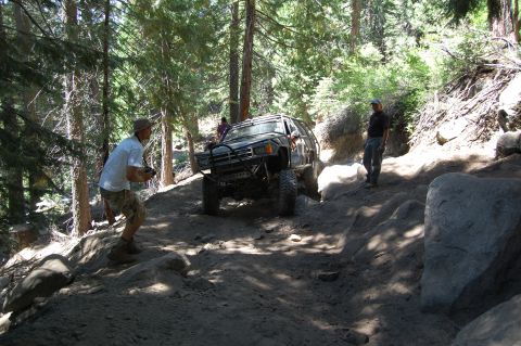 1993 Jeep Wrangler - A modified off-road vehicle tackles a challenging, wooded trail with large rocks and steep inclines.