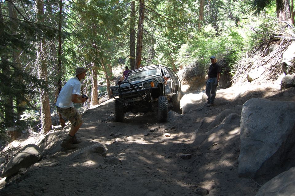 Off-road vehicle navigating a steep, rocky trail with trees on either side. A person stands nearby filming