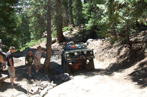 1993 Jeep Wrangler - People watch an off-road vehicle navigate a rocky trail in a wooded area.