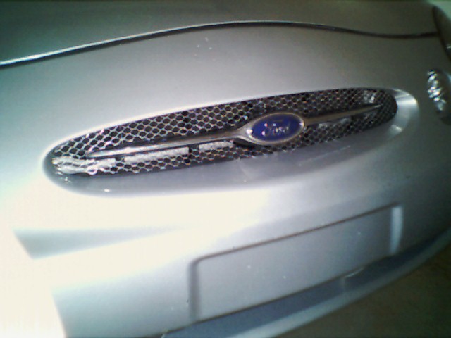 Silver Ford Taurus grille with blue oval Ford badge and honeycomb mesh. Visible portion of bumper below