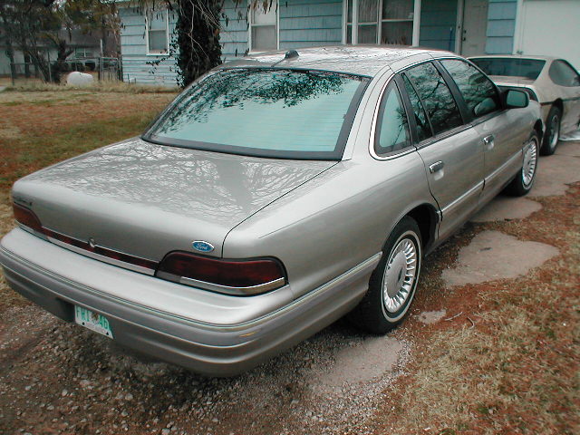 Silver Ford Crown Victoria sedan, rear three-quarter view, parked on gravel driveway by a blue house