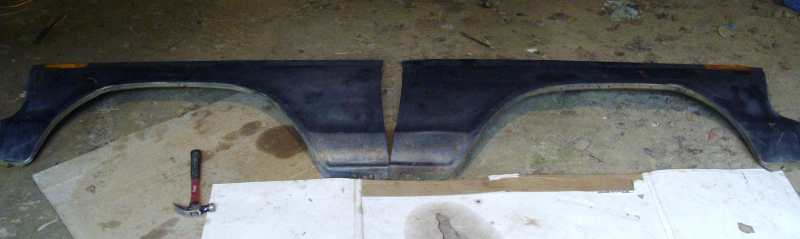Two dark blue fender flares laid out on the ground with a hammer nearby. Supermotors.net logo in the corner