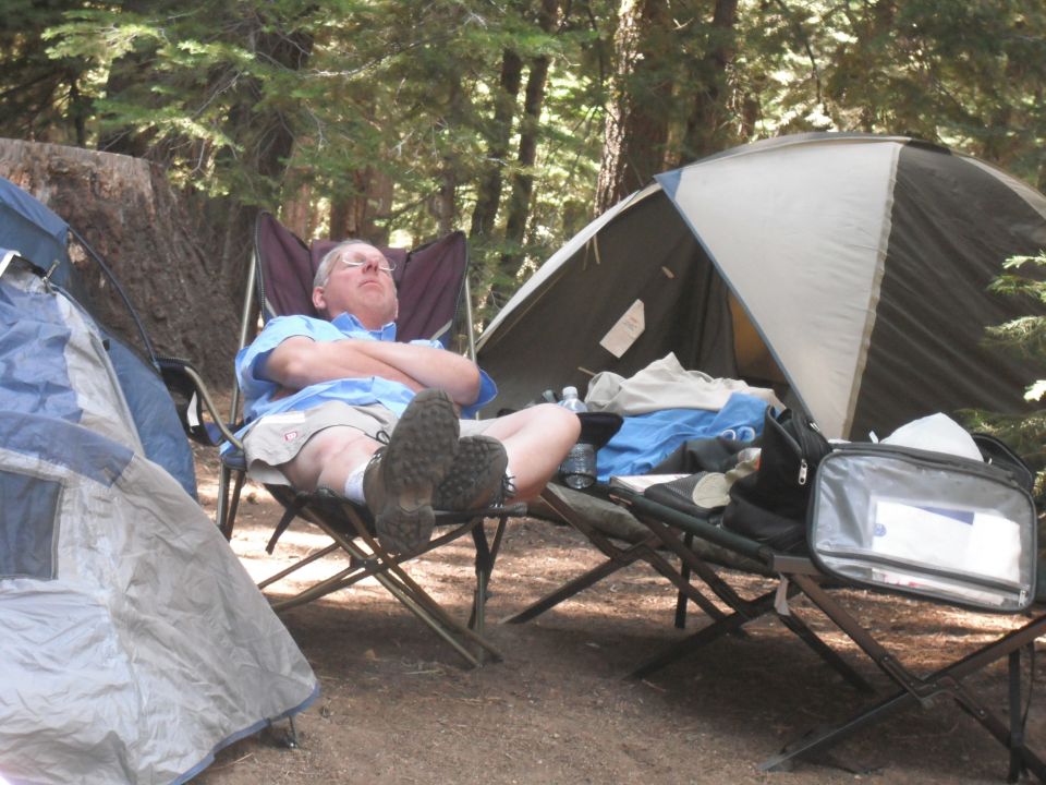 Man sleeping in a camping chair at a campsite with tents and trees