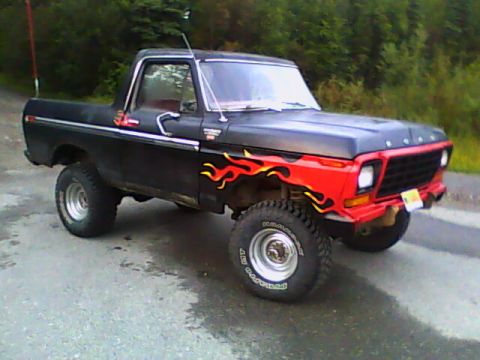 1978 Ford Bronco - A black 1978 Ford Bronco with flame decals is parked on a paved surface with trees in the background.